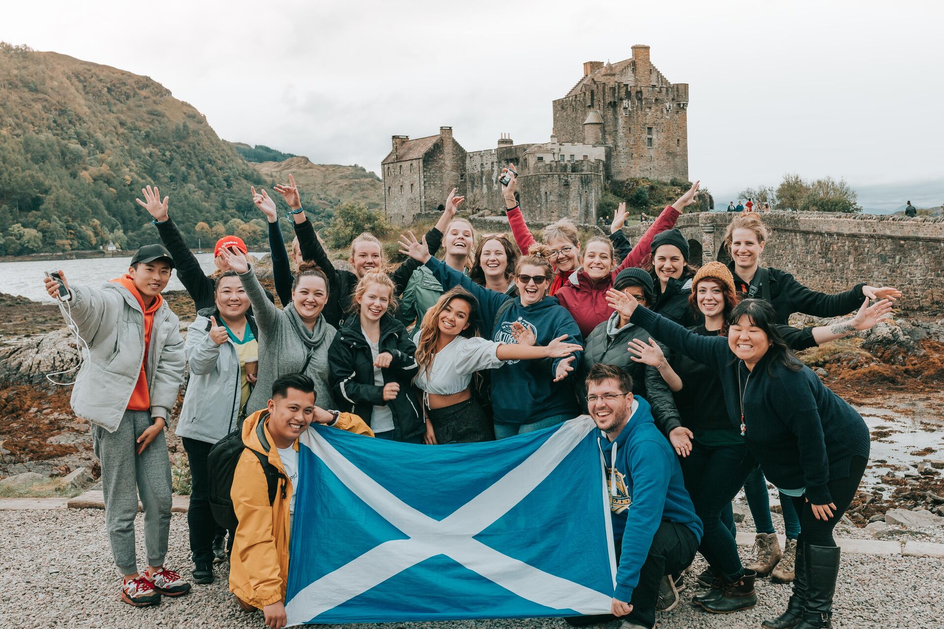Large group of young travellers holding a Scottish flag in front of Eilean Donan Castle