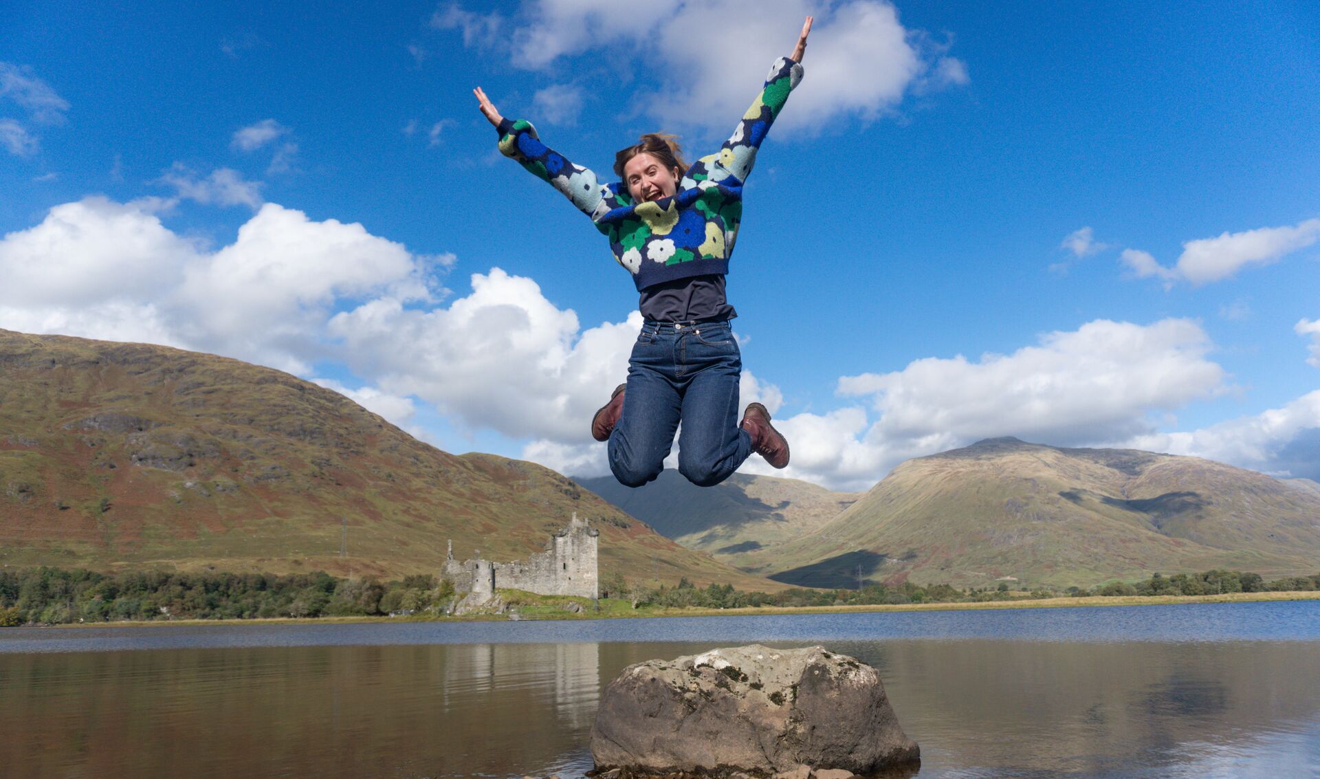 Young woman jumping with sunny views of Kilchurn Castle across the shore
