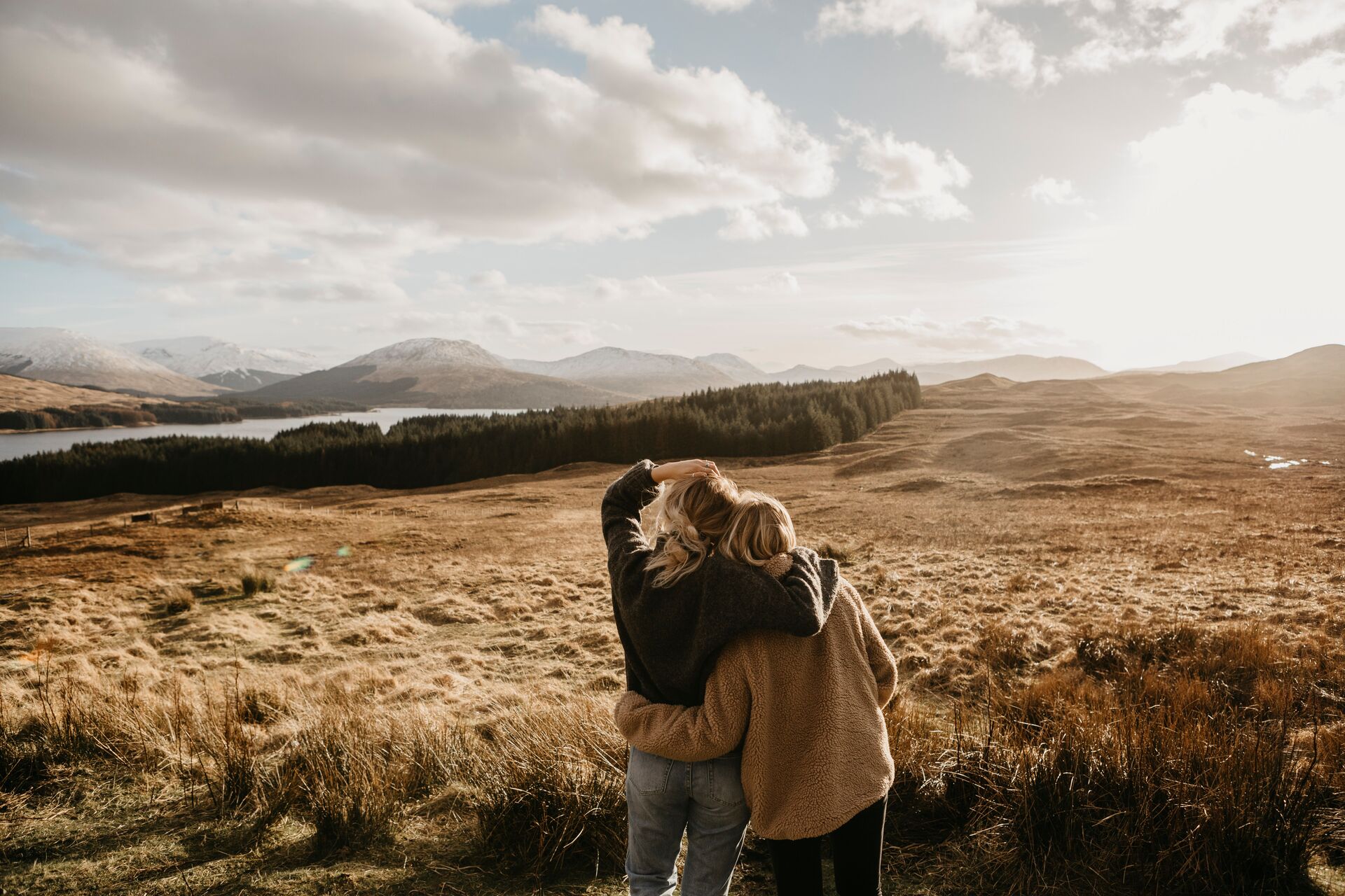 Two young women embracing while watching the sunset over vast open land with distant hills and forests